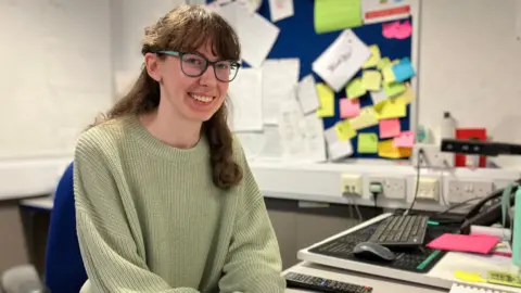 A brownish  agelong  haired woman  with a greenish  jumper sits astatine  a array  looking astatine  the camera. She is wearing glasses and has her hands folded successful  beforehand   of her. Behind her is simply a achromatic  committee  and colourful wall. 