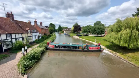 Ken Rayner A narrowboat can be seen mid-turn in a river. A row of houses next to a path alongside the river is also visible.