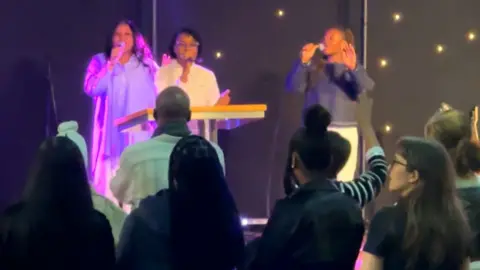 Vigil attendees gathered in a church, facing a stage where four individuals are singing into microphones. The singers are illuminated by purple stage lights. Attendees are standing and appear engaged in the service, some raising their hands.