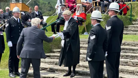 A petitioner wearing a grey suit hands over a document to the clerk, who is wearing a black ceremonial gown and wig.