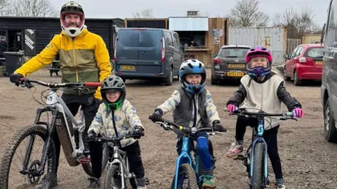 Contributed Ryan Horsfall and his three young children who all sit on bikes and wear helmets. They are smiling at the camera. 