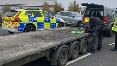 Cambridgeshire Police The rear end of a flat bed trailer side-on, which has three tyres at its rear. A man in black is bending over its rear. Behind it is a side view of a yellow and blue police car