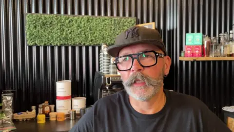 A man with a moustache and glasses stands in an a cool shop that sells and rents E bikes.