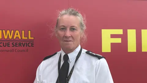 Kathryn Billing smiling and stood in front of a red fire engine. She is wearing a white shirt, with a black tie. 