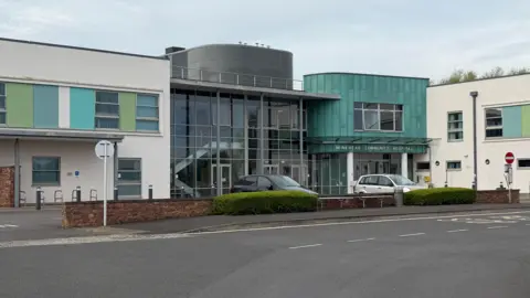 Exterior of the community hospital, with a green facade over the entrance, and window wall looking inside. A few cars are outside.