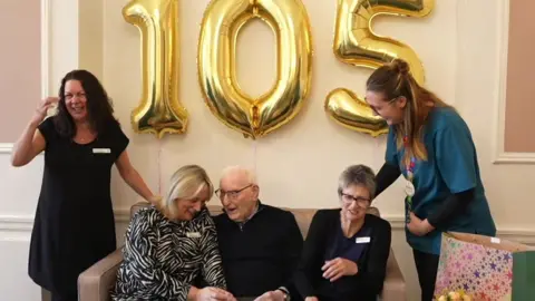 Ron sits on a sofa animatedly chatting to a woman on his right. The women around him, some of whom are care home workers, are all laughing. There is a gold 105 balloon behind them.