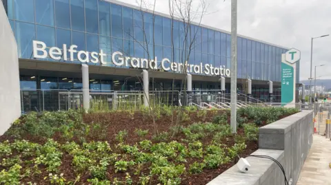 The front of Belfast's Grand Central Station, a large glass-fronted building with a large white entrance sign. There is a new flowerbed enclosed behind the exterior wall of the station. 