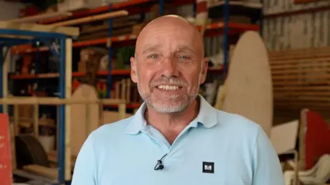 BBC A bald man smiles at the camera, and he is wearing a pale blue polo shirt. In the background there are pieces of wood to build a float for the Battle of Flowers parade. 