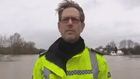 Mark Sitton-Kent with brown hair and red-framed glasses, wearing a high vis jacket and stood in front of a raised river.
