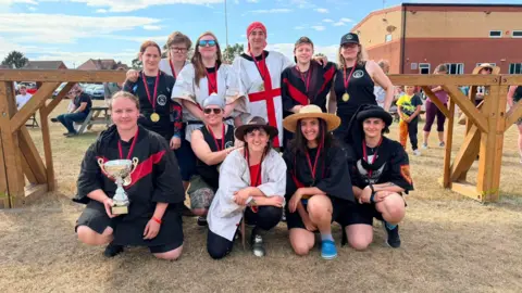 Dave Polier. Diez mujeres sonriendo para la cámara. Una sostiene un trofeo de campeona y todas tienen medallas. Algunas aún visten atuendos que recuerdan al combate medieval, mientras que otras llevan ropa deportiva moderna.