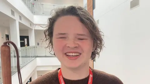 Annabel Amos/ BBC A girl with long brown hair wearing a brown jumper and a red University of Northampton lanyard stands in a stairwell. 