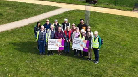 A crowd of people huddle together holding up two signs that read "National Walking Month".