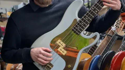 PA Media A man wearing a navy blue jumper holding a bass guitar. The guitar is white with a gold inlay.
