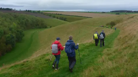 Pockington Ramblers Ramblers spread over a long distance on a green grass path on a hillside. To the left of the picture the grass steeply slopes down to a patch of woodland