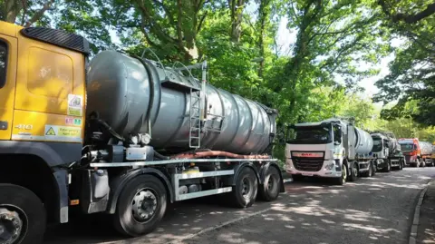 Tankers nose to tail along a tree-lined road.
