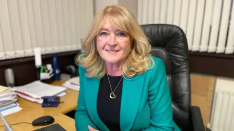 Geraldine has blonde shoulder-length hair, blue eyes and is smiling. Sitting in a black office chair at her desk, she is wearing a turquoise blazer over a black t-shirt, and around her neck a small gold heart hangs from a thin gold chain.