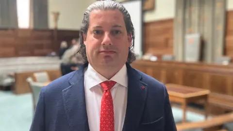 Owen Sennitt/BBC A man with slicked back hair wearing a suit, white shirt and red tie looks towards a camera in a council chambers