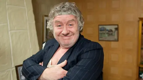 Gregor Fisher as Rab C Nesbitt. He is wearing a curly wig, black pinstripe blazer and string vest.