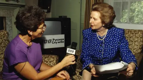 Two women, one with a microphone speaking to a woman in a blue blouse