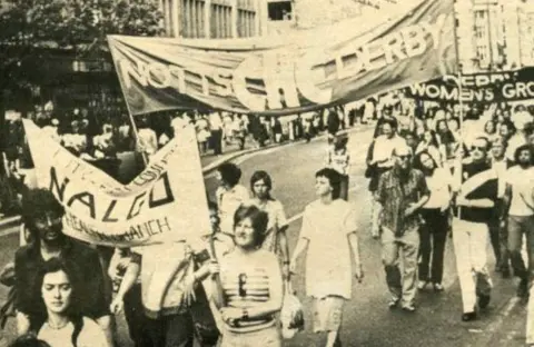 Rainbow Heritage A black and white image of women and men marching with banners reading 'Notts CHE Derby'. There are three banners in the photo. It looks like a summers day with people wearing sunglasses.