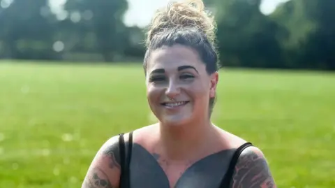 A smiling woman with heavy tattoos sits on some grass.