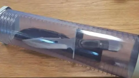 PSNI A hunting knife is placed into a plastic container, which is on a wooden table.