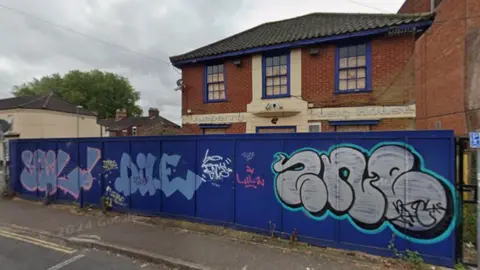 The outside of the former Blueberry Music House in Cowgate in Norwich. The building has blue window frames which stands out against the brown bricks and lettering is still attached to the front of the building, which reads Blueberry Music House. In front of the building is a stretch of blue boards covered in graffiti.