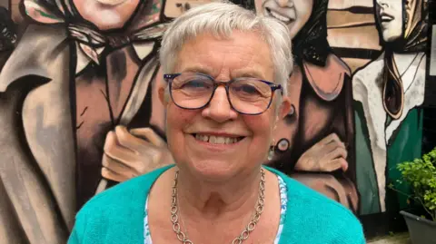 Gabrielle Deans has short white hair and is wearing a light blue top and silver necklace. She has black-rimmed glasses and is seen smiling. She is standing in front of the factory girls mural in Derry.