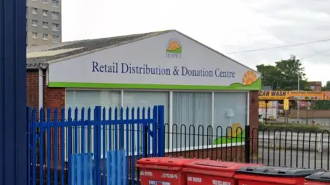 The exterior of Lindsey Lodge Hospice retail distribution and donation centre. It is a brick building with a white and green charity logo and "Lindsey Lodge Hospice Retail Distribution & Donation Centre" written in blue writing on the front apex. Below that is four large windows. The building sits behind black and blue metal railings. Red bins sit in front of the railings. The sky is cloudy.