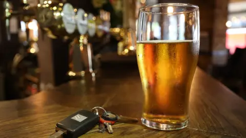 A set of car keys next to a pint of beer on a pub bar. The beer is only two thirds full.