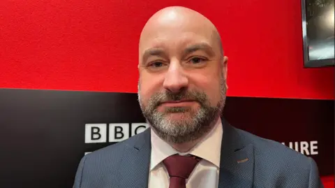 Marc Jones stares into the camera. He has a bald head and a greying beard. Mr Jones is wearing a blue suit, maroon tie and a white shirt. He is in a radio studio with red walls and a black logo.