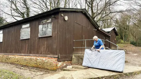 Tansley Wood Scout Campsite Two large wooden huts with a brick base, three windows on one side and large double doors. A woman is stood outside inspecting a striped mattress which has a large whole in it. The woman has blonde hair, glasses, a blue shirt and scout neckerchief. There are lots of trees behind the huts.
