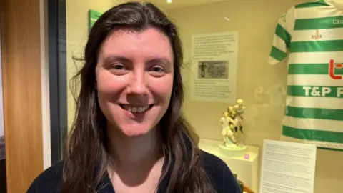 BBC/Claudia Robinson A woman with long brown hair smiles at the camera, with a small statue in the background in a glass cabinet, alongside a football shirt.