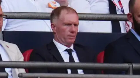 Reuters Paul Scholes is sitting in the stands at Salford City football club. He has light ginger hair and is wearing a black jacket and black tie with a white shirt.