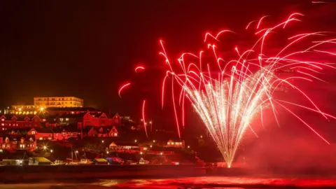 A red firework set off from the coast. There are houses on a hill. It is nigh time. The sea is lit up red from the firework. The sky is black.