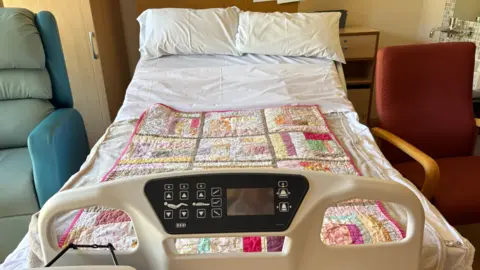 A picture of the 'cuddlebed' from the bottom end looking towards the pillows in at the far end. It has a digital display and controls to adjust the bed, with a coloured blanket on the white bedspread. 