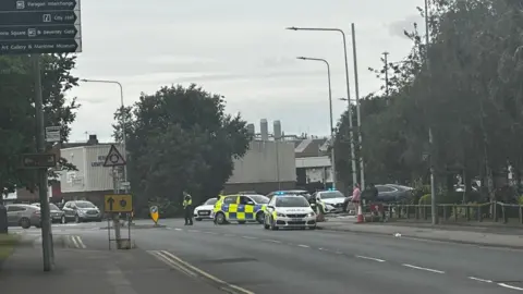 Amber Gash/BBC Three police cars block the entrance to a car park. A number of industrial buildings are visible in the background. A row of trees is lining the road.