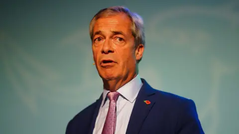 Nigel Farage stood in front of a Welsh dragon in Reform colours, wearing a blue suit, shiny purple tie and purple shirt. He has a microphone and a small poppy.