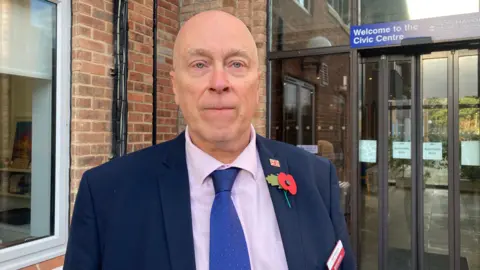 Councillor Rob Wharton is looking at the camera while standing outside a council building, wearing a blue suit and pink shirt. Behind him on the right is a glass-doored entrance with a sign saying "Welcome to the Civic Centre" above it.