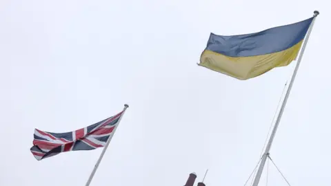 A Union Jack and Ukrainian flag flying side by side in front of a grey sky.