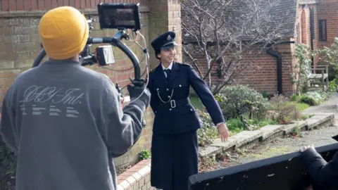 A woman acting as a police officer being filmed by a man in a yellow beanie with a big camera attached to a right. The actress is smiling and walking along the road.