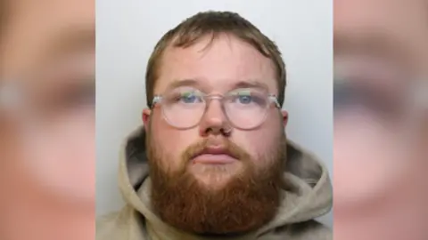 A police custody image of a man with ginger hair and a large bear wearing a hoody and glasses. 