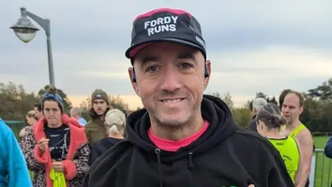 A man with a black top and black cap among a crowd of people in running tops in a park with metal railings behind him