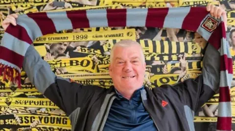 Grant Danskine, holding the claret and blue scarf above his head and standing in front of a poster of football team Young Boys which features lots of yellow and black scarfs