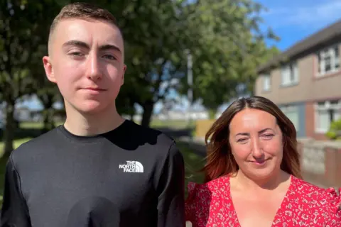 Ryan, wearing his vacuum bell under his t-shirt, stands beside his mum Liza - who has long brown hair and is wearing a red floral dress. They are outside their home, with trees in the background.