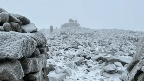 Stephen Oliver The rocky mountain top is dusted with white snow. A group of people standing on a rock at the summit can just be made out through the fog. They are viewed from a position lower down, with frosty rocks closer in view to the left. The route up to the summit is strewn with stones and rocks, in shades of grey and white from the light dusting of ice and snow.