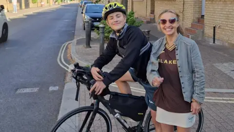 Victoria Bacon Ranulph, wearing a full-sleeve navy shirt, navy shorts and a yellow helmet, sitting on a cycle. His mum is standing next to him, and is wearing a brown shirt, grey jacket and white skirt. The pair are in a street, smiling for the camera. 
