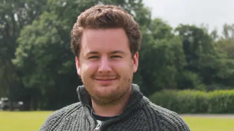 A close-up of Thom Campion smiling as he looks directly into the camera. He has brown hair, brushed away from his face, and a thin beard. He is wearing agreen woolly top and standing in a field with trees behind him.