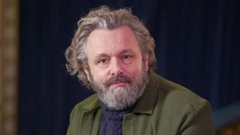 BBC Michael Sheen ha la barba grigia e i capelli lunghi. Indossa un maglione lavorato a maglia blu e una giacca verde