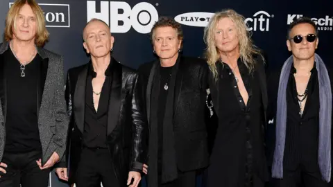 Getty Images Joe Elliott, Phil Collen, Rick Allen, Rick Savage e Vivian Campbell, da Def Leppard, participam da cerimônia de indução do Hall da Fama do Rock & Roll 2019. Existem logotipos atrás deles no cenário. Os homens estão vestidos de preto e Vivian Campbell tem um lenço cinza.